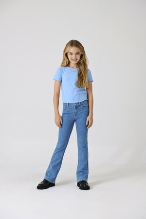 Joven chica de pie con postura relajada, vistiendo una camiseta de manga corta azul claro, vaqueros azules y zapatos negros sobre un fondo liso.