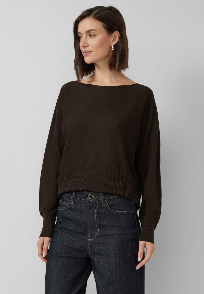 Femme aux cheveux bruns courts portant des boucles d'oreilles en argent, un pull brun foncé et un jean bleu foncé, regardant vers sa gauche sur un fond uni.