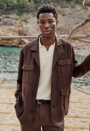 Ung mand iført en brun jakke og bukser med en hvid skjorte, står ved et trægelænder nær vand og klippefyldt terræn.