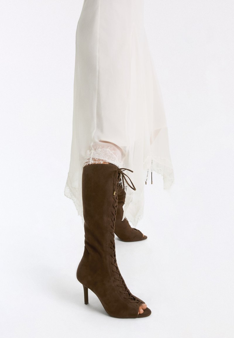 Bottes en daim marron montantes jusqu'au genou à lacets et bout ouvert, portées avec une robe blanche bordée de dentelle sur fond blanc uni.