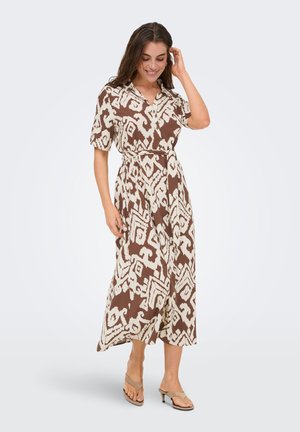 Femme portant une robe midi à motifs marron et blanc avec des manches courtes et des sandales beiges à talons, souriante et touchant ses cheveux.