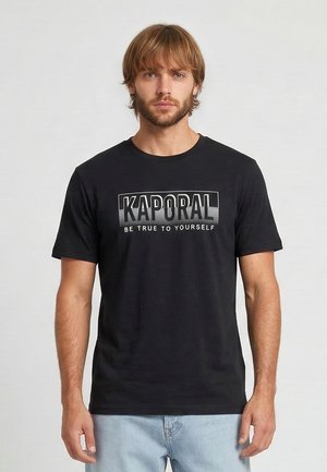 Homme aux cheveux châtain clair et barbe, portant un t-shirt noir Kaporal avec le texte « Be true to yourself », debout devant un fond clair uni.