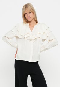 Licht crèmekleurige blouse met gelaagde ruches over de borst, gestructureerde kraag, lange mouwen en een knoopsluiting aan de voorkant.
