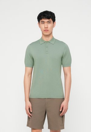 Giovane uomo indossa una polo verde salvia a maniche corte e pantaloncini cargo marroni, in piedi davanti a uno sfondo bianco uniforme.