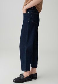 Donkerblauwe denim jeans met een relaxed fit, taps toelopende pijpen en een traditionele vijf-pocket ontwerp. Gemaakt met zwarte instappers en een beige top.