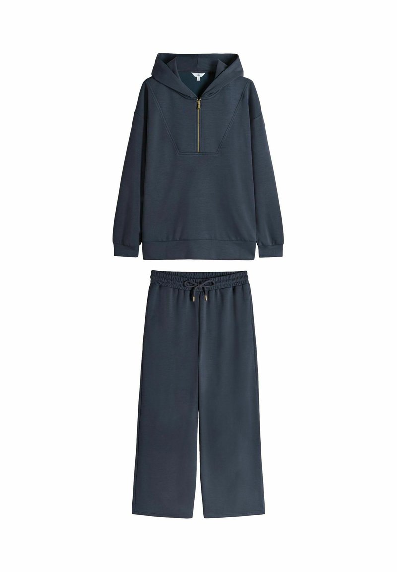 Felpa blu navy con mezza zip, vestibilità oversize e tasca a marsupio, abbinata a pantaloni larghi coordinati con cintura elastica.