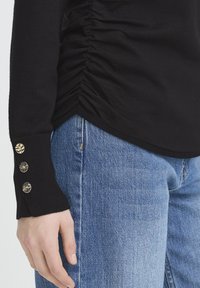 Czarna bluzka z długim rękawem z marszczoną fakturą po bokach; ozdobiona złotymi guzikami przy mankiecie. Zestawiona z niebieskimi dżinsami.