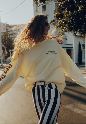 Femme aux longs cheveux bouclés portant des lunettes de soleil, un sweat-shirt jaune et un pantalon à rayures noires et blanches, marchant en extérieur par une journée ensoleillée.
