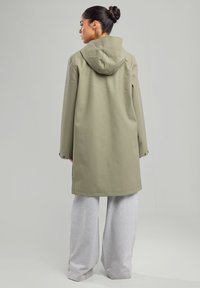 Khaki veden säältä suojaava takki, jossa on huppu, pitkät hihat ja napit suljin hihoissa, yhdistettynä vaaleanharmaisiin väljät sweatpants-housuihin.