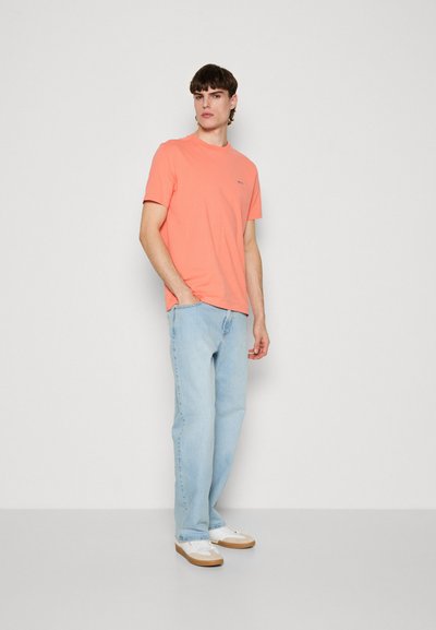 T-shirt corail à manches courtes, coupe décontractée, associé à un jean bleu clair. Des baskets blanches avec une semelle marron ajoutent un contraste décontracté. Design minimaliste.