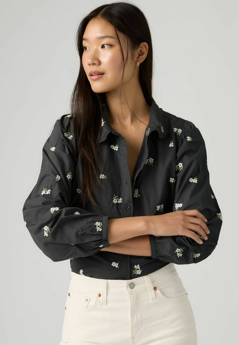 Chemise noire boutonnée avec broderie florale en blanc et vert. Caractérisée par des manches bouffantes et un col, portée avec un pantalon blanc.