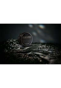 Schwarzer runder Behälter des "Snail Bee Ultimate Hydrogel Eye Patch" mit weißem Text und glatter Oberfläche, vor einem dunklen, steinigen Hintergrund platziert.
