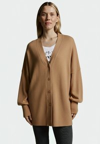 Beige Cardigan aus strukturiertem Stoff, mit V-Ausschnitt, oversized Fit und fünf Frontknöpfen. Über einem weißen Oberteil getragen.