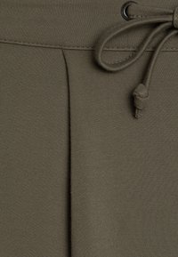 Pantaloni jogger verde oliva realizzati in un morbido tessuto elasticizzato. Presentano una vita regolabile con cordino e un delicato dettaglio a piega. Texture liscia.