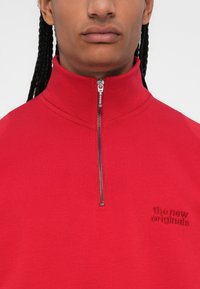 Suéter rojo de cremallera con cuello alto, textura acanalada y un pequeño logo bordado en el lado izquierdo del pecho. La cremallera tiene un herraje metálico.