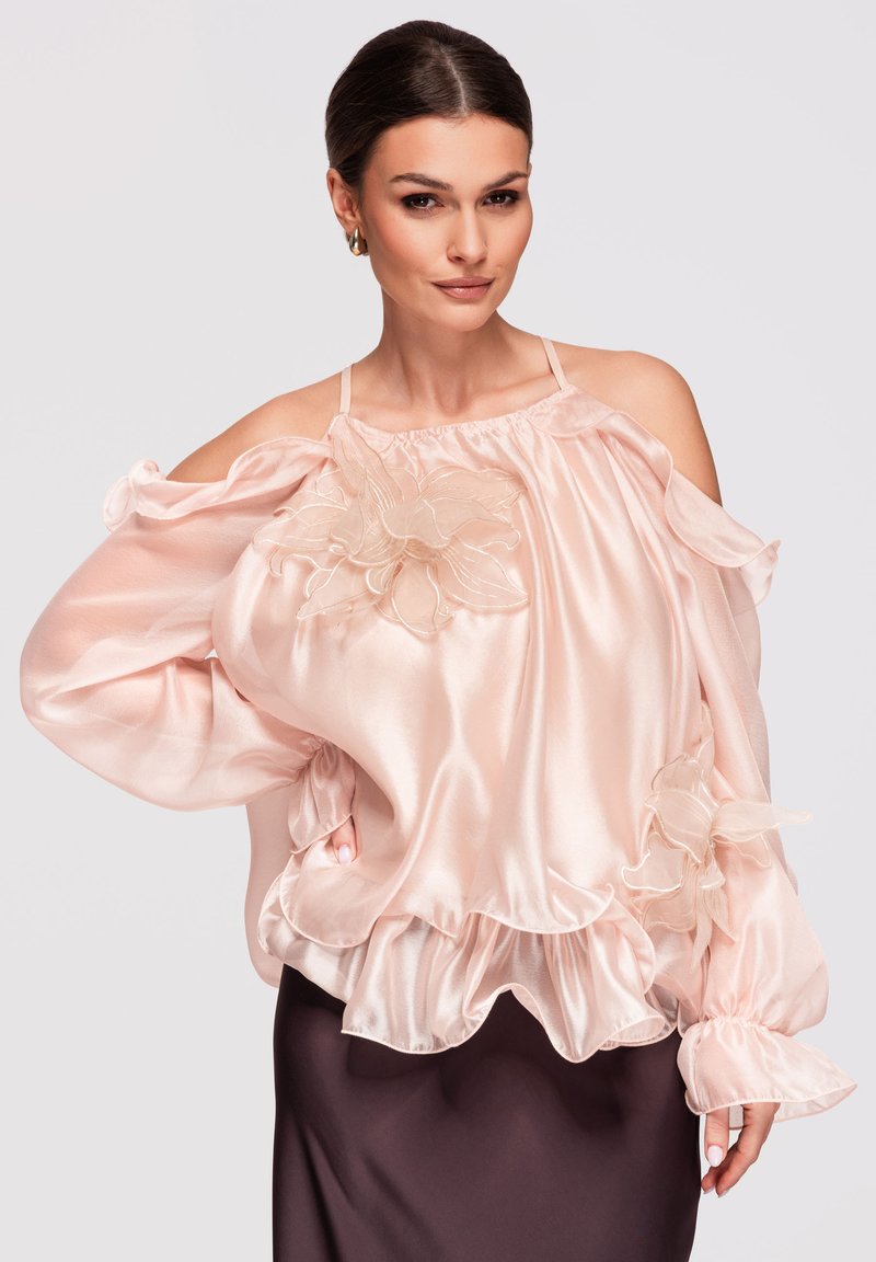 Femme portant une blouse en satin rose pâle épaules dénudées avec des appliqués floraux et un ourlet à volants, associée à une jupe foncée.