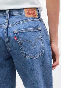 Κοντινό πλάνο της πίσω τσέπης μπλε τζιν Levi's, που δείχνει το δερμάτινο ετικέτα της μάρκας και την κόκκινη ετικέτα Levi's, φορεμένα από άτομο με λευκό πουκάμισο.