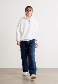 Sweat à zip blanc avec un col montant, associé à un jean large bleu et des baskets blanches, se tenant devant un fond neutre.