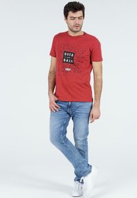 Czerwona bawełniana koszulka z nadrukiem graficznym "OVER AND BACK", w połączeniu z jasnoniebieskimi dżinsami i białymi trampkami. Prosty design, codzienny krój.
