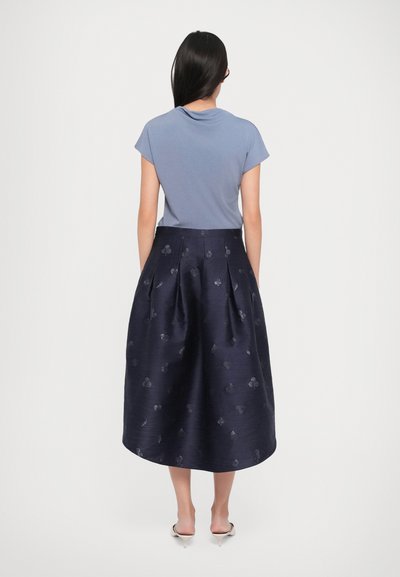 Woman with long black hair wearing a blue short-sleeve top, navy midi skirt with subtle club and spade patterns, and white heeled sandals.