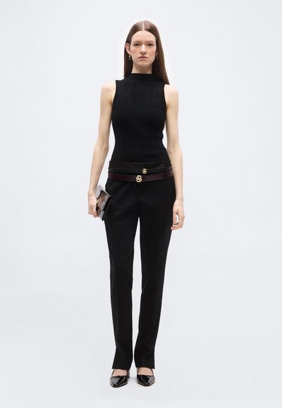 Femme portant un haut noir sans manches, un pantalon droit noir, deux ceintures noires et brun foncé avec des boucles dorées, des talons noirs, et tenant une pochette noire.