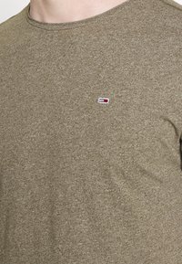 Detailní záběr na osobu, která má na sobě texturované olivově zelené tričko s malým obdélníkovým logem v červené, bílé a modré barvě na levé hrudi.