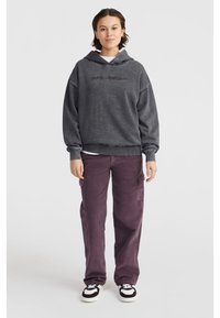 Felpa grigia con cappuccio e logo frontale, abbinata a pantaloni cargo viola larghi e sneakers in bianco e nero.