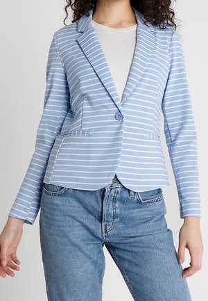 Mujer con una blazer de rayas azul claro y blanca sobre una camisa blanca con vaqueros azules contra un fondo liso.