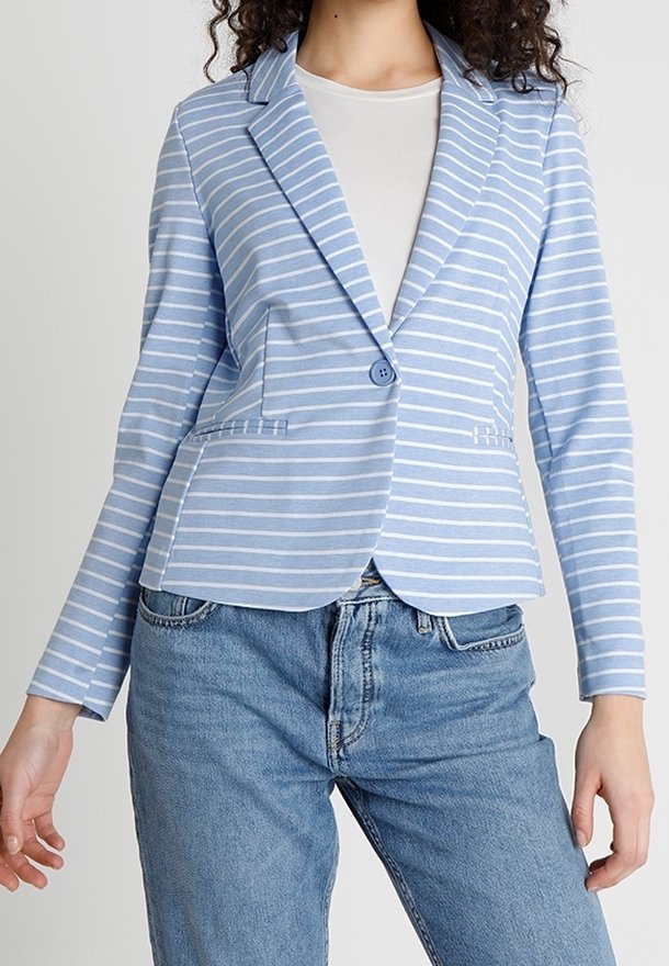 Femme portant un blazer rayé bleu clair et blanc par-dessus une chemise blanche avec un jean en denim bleu sur un fond uni.