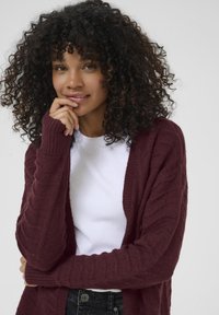 Cardigan tricoté bordeaux avec un motif texturé, à manches longues et à ouverture devant, porté par-dessus un haut blanc. Un pantalon noir est visible au niveau de la taille.