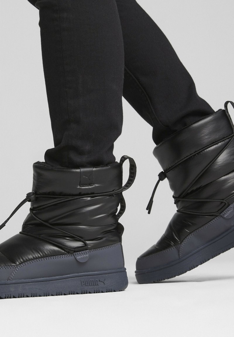 Bottes d'hiver noires et rembourrées avec une tige haute, dotées d'un système de laçage ajustable et d'une semelle en caoutchouc texturée. Marque visible sur le côté.