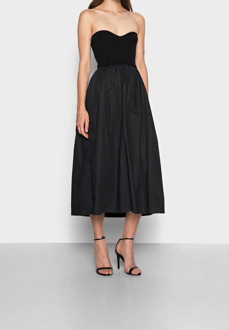 Femme portant une robe midi noire bustier avec un corsage ajusté et une jupe ample, associée à des sandales noires à talons hauts et bride à la cheville.