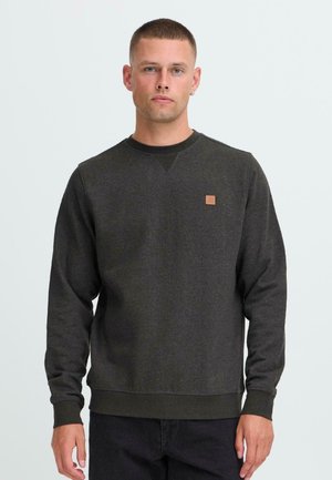 Jonge man met kort haar, gekleed in een donkergrijze ronde hals trui en zwarte broek, staand tegen een effen lichte achtergrond.