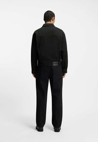 Musta farkkutakki, jossa on jäsennelty muotoilu ja nepparit, yhdistettynä mustiin housuihin. HUGO-logo takin selässä.