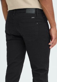 Crni traperice s uskim krojem, dvije stražnje džepove i teksturiranim oznakom na struku. Tkanina izgleda glatko s laganim rastezanjem.