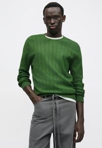 Zielony prążkowany sweter z okrągłym dekoltem, w połączeniu z szarymi casualowymi spodniami z wiązaniem w pasie. Gładka faktura, luźny krój.