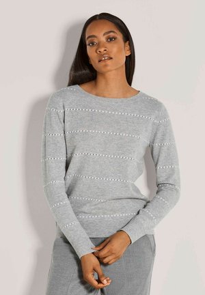 Frau mit langen dunklen Haaren, die einen hellgrauen Strickpullover mit horizontalen Lochstreifen und passenden grauen Hosen vor einem schlichten Hintergrund trägt.