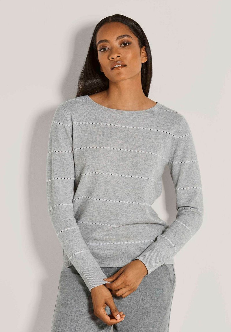 Frau mit langen dunklen Haaren, die einen hellgrauen Strickpullover mit horizontalen Lochstreifen und passenden grauen Hosen vor einem schlichten Hintergrund trägt.
