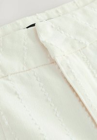 Bijela tkanina s teksturiranim uzorkom i blagim rebrastim učinkom. Ima presvučen pojas i detalje s nevidljivim šavovima.