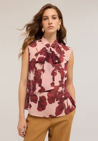 Donna con capelli mossi che indossa una blusa senza maniche di colore rosa con una grande stampa floreale rossa e un dettaglio a fiocco, abbinata a pantaloni color senape.