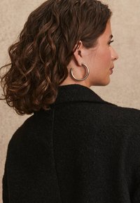 Schwarzer strukturierten Mantel mit einem Schalkragen, getragen über einer lockigen Frisur. Das Modell trägt große Creolen, die den Ohr- und Halsbereich betonen.