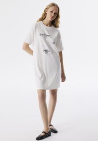 Witte katoenen T-shirt jurk met korte mouwen. Heeft zwarte grafische prints op de voorkant, een ontspannen pasvorm en een knie-lange lengte. Zwarte platte schoenen.