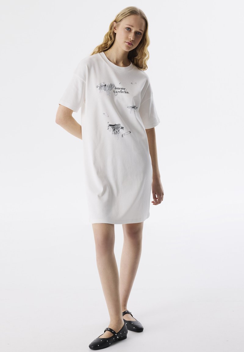 Vestido de camiseta de algodón blanco con mangas cortas. Presenta estampados gráficos en negro en la parte frontal, corte relajado y diseño hasta la rodilla. Zapatos planos negros.