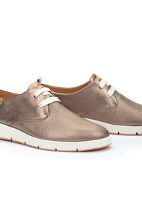 Zapatillas metálico bronce con textura perforada, cordones blancos y suela blanca. Presentan un acabado suave similar al cuero y un diseño minimalista.