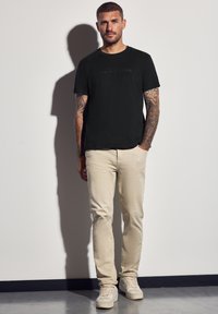 Muž s tetováním, oblečený v černém tričku, béžových kalhotách a teniskách, stojící před bílou zdí s předpokládaným stínem za sebou.