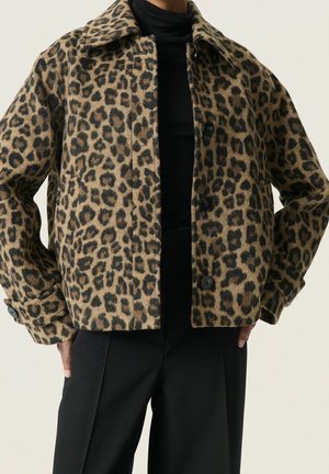 Donna con giacca corta a stampa leopardata sopra un dolcevita nero e pantaloni neri, mani posate sui fianchi su sfondo uniforme.