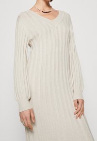 Beige geribde gebreide jurk met een v-hals, lange pofmouwen en aansluitende manchetten. Zachte textuur met verticale strepen door de gehele jurk.