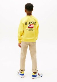 Felpa gialla con logo ricamato e dettaglio a righe sul collo. Pantaloni beige e sneakers multicolore completano l'outfit.