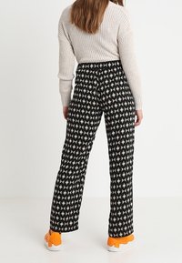 Svarta mönstrade byxor med diamandmönster, med elastisk midja och vida ben. Bärs tillsammans med en ljus tröja och orange skor.