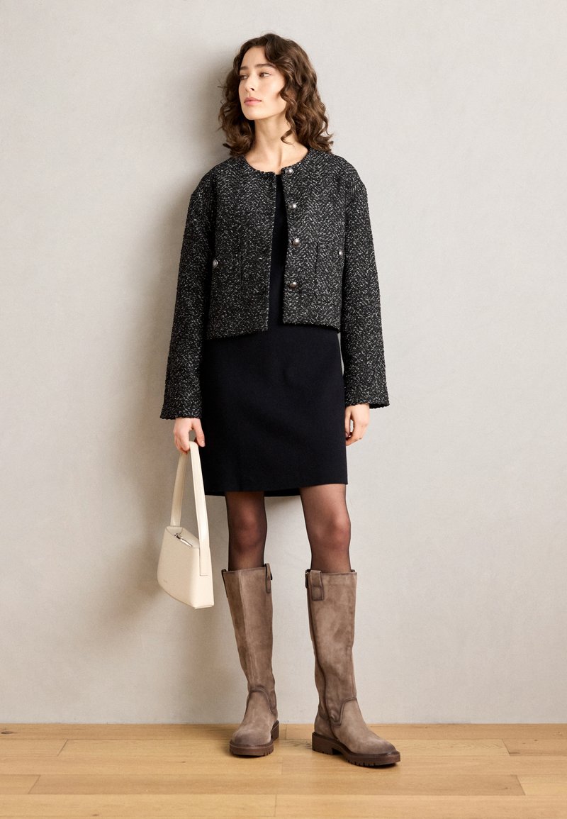 Veste cropped en tweed noir et blanc avec un col rond, de grands boutons, portée avec une robe noire, des bottes en suède marron à hauteur de genou et un sac beige.